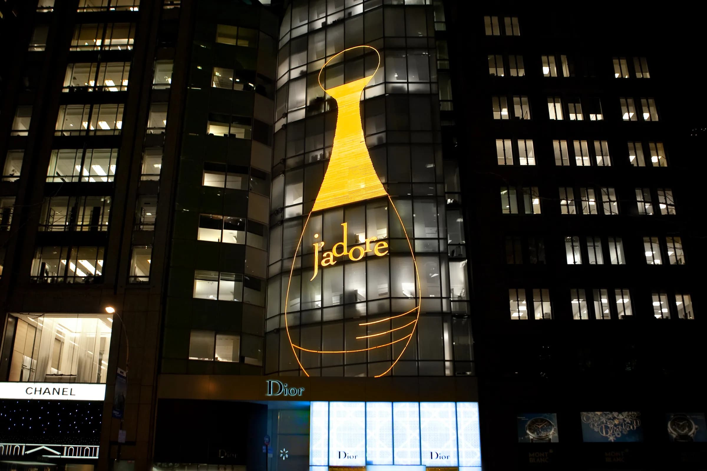 Dior 57th Street flagship at night, its glass tower outlined with a glowing gold J'adore bottle, New York